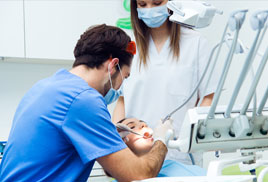 hombre y mujer de profesión odontólogos, atendiendo a una mujer en el consultorio, estan vestidos con ropa especial de odontología color blanco